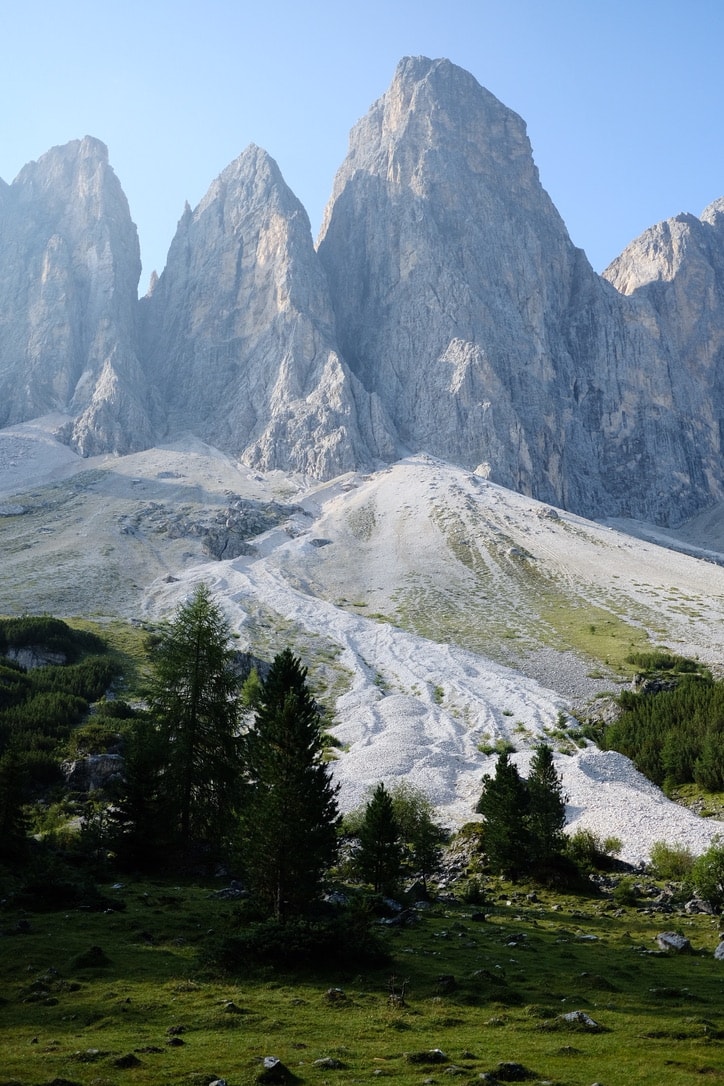 The Geisler massif seen in portrait, a pale scree fan running down toward spruce and larch trees on green pasture