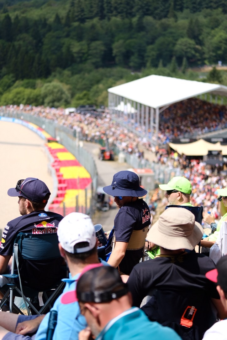 Spectators in caps and bucket hats on a grass bank in the foreground, looking down at a race track with red and yellow kerbs and packed grandstands and forest behind it