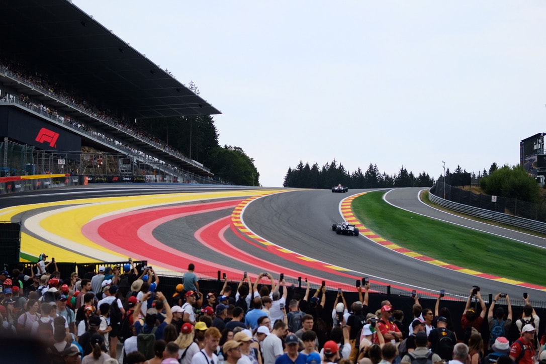 A wide sweep of race track lined with red and yellow kerbs, a packed grandstand on the left and a row of spectators along the fence in the foreground holding up phones as two cars go by