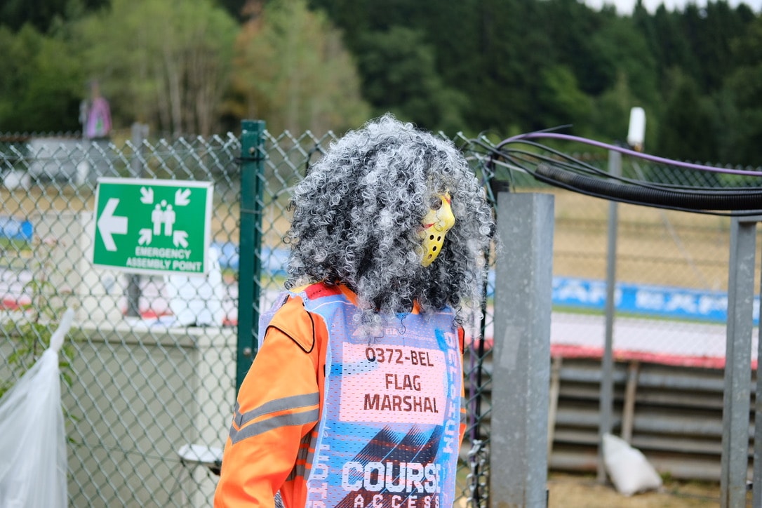 A flag marshal seen from behind in orange overalls and a course access tabard reading 0372-BEL FLAG MARSHAL, with long grey curly hair and a hockey mask visible in profile, standing at a chain-link fence beside an emergency assembly point sign
