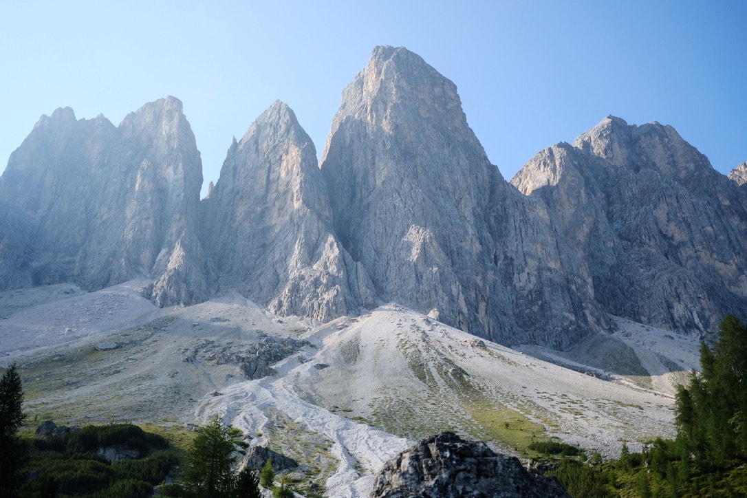 The Geisler peaks in morning light above pale scree slopes, with larches and spruce along the bottom of the frame