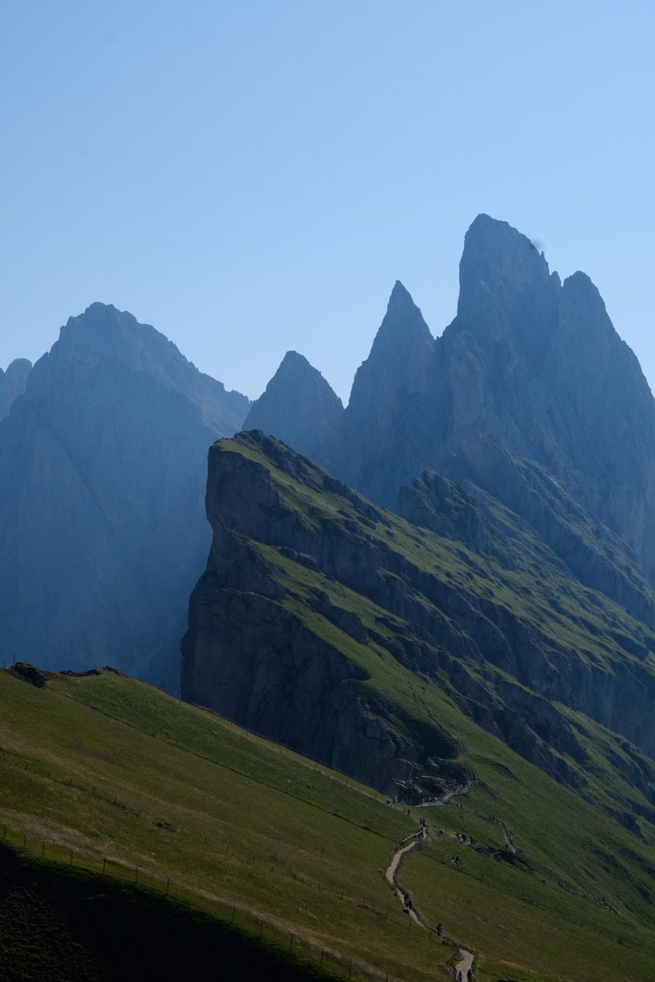A narrow trail traversing the grassy flank of the Seceda ridge, with layered blue rock spires stacked behind it