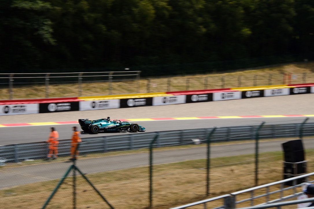 A green Formula 1 car blurred by motion as it passes a barrier of sponsor boards, with two marshals in orange overalls standing trackside behind it