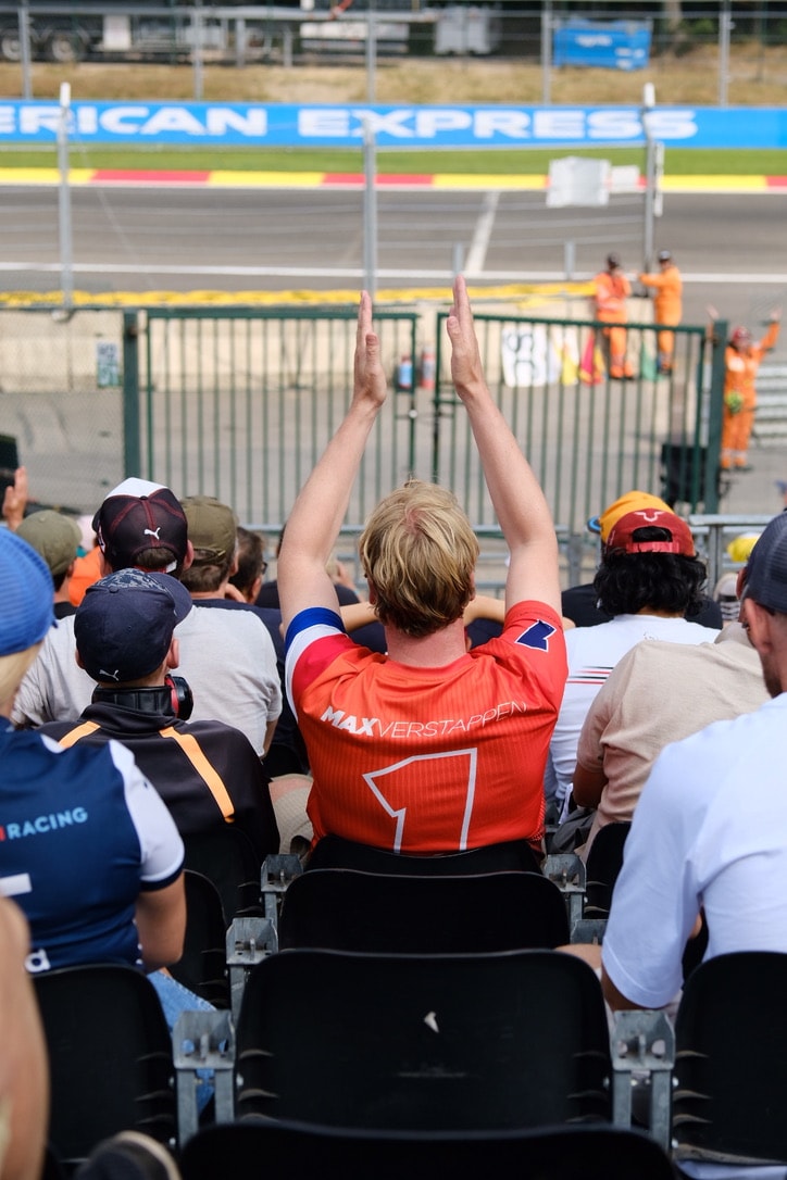 A spectator seen from behind in an orange Verstappen number one shirt, both arms raised, standing in a grandstand above the track with marshals in orange on the far side